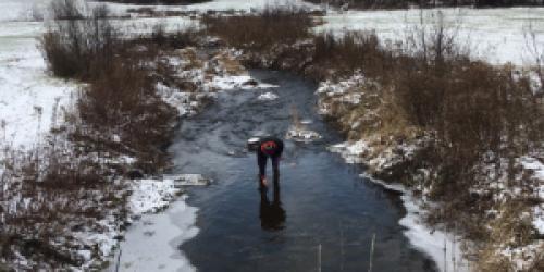 person dipping a sample vial in a stream during winter