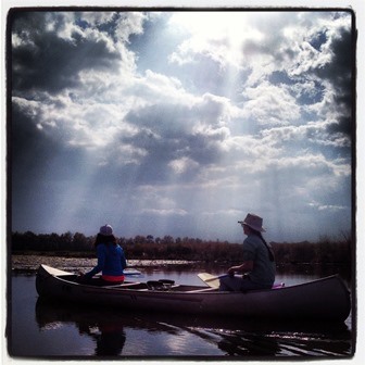 Paddling in Riverine Wetlands.  Two people are seen on a canoe paddling across water.  They are not wearing lifejackets and are partially silhouetted on the bright light coming from the sun.  The sky is partly cloudy with rays of sunshine shining through the clouds.