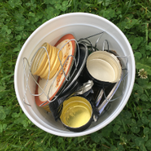 An empty yogurt container holding small scrap metal, including bottle caps, wire, jar lids, a broken carabiner, and more.
