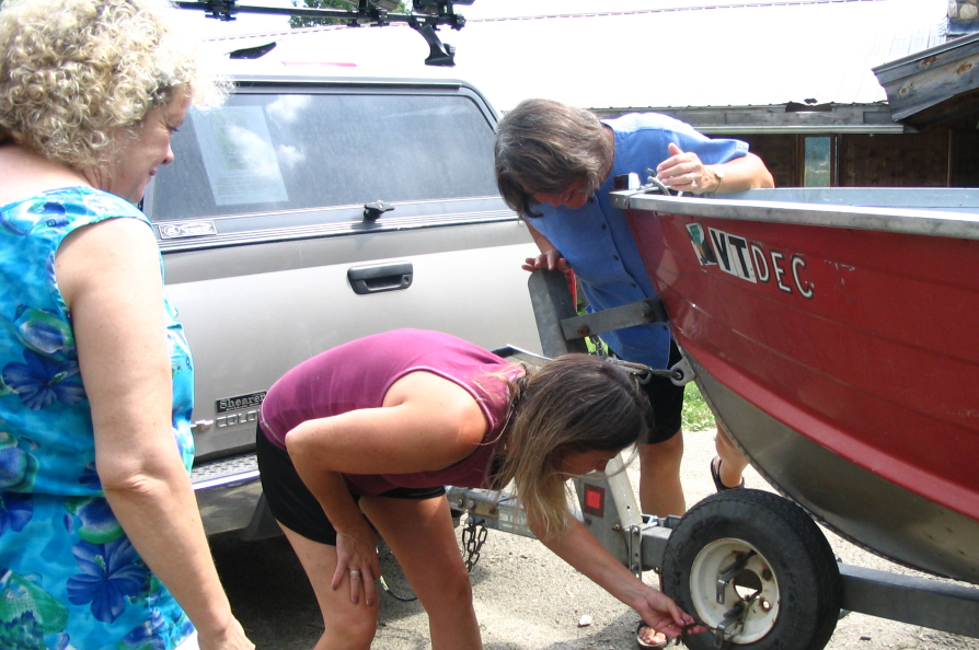 A women in the center bends forward and inspects a boat trailer with a red boat on it as two other people watch from both sides.