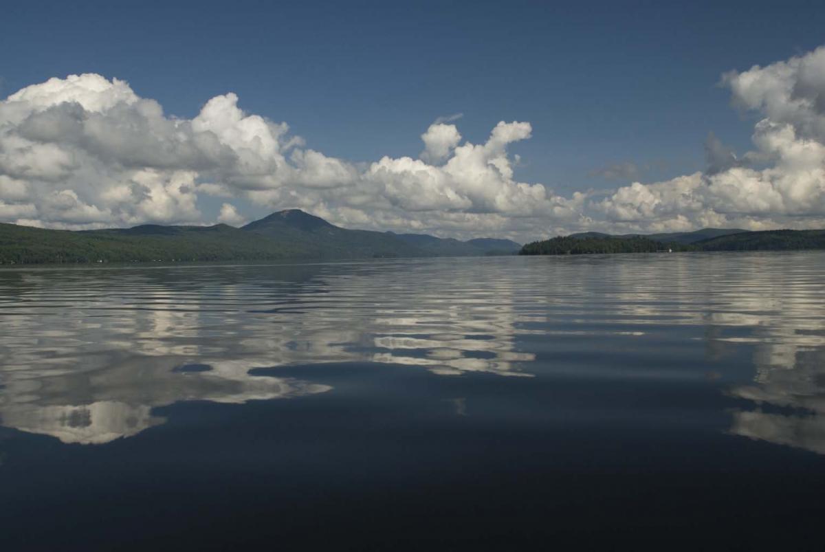 A view of Lake Memphremagog from the south looking north.  Owl's Head Mountain is seen in the background.  The sky is blue with some puffy clouds and the vegetation on the distant shore is green.