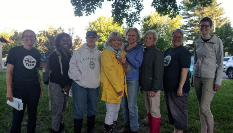 Eight people standing outdoors in the shade on a sunny day facing the camera smiling.