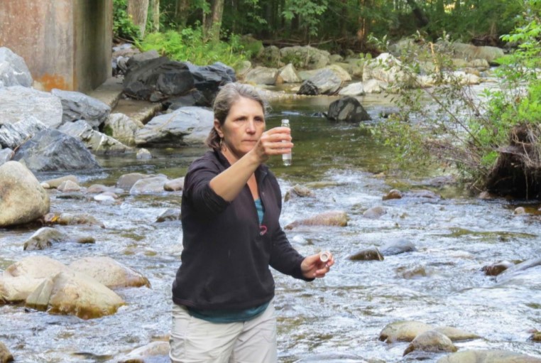 A woman wearing a black sweatshirt standing in the middle of a shallow rocky stream holding up and looking at a vial of water