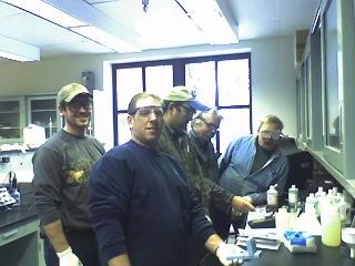 Five people working in a lab.  They are all wearing safety glasses and on the right there is a table with bottles and small instruments on it.