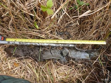 Tape measure indicating depth of sample taken for soil profile.  A yellow tape measure is seen next to a grey metal rod which is next to some grey soil.  All is laid on top of brown grassy material.