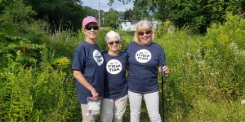 three people standing by a stream wearing Stream Team shirts and holding water sampling equipment